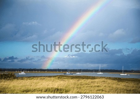 A vivid rainbow arcs from dramatic storm clouds over a sheltered coastal harbour dotted with moored yachts, fringed by golden saltmarsh. Atmospheric British coastal landscape.