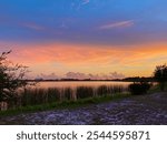 A vivid sunset over calm waters in Gulf Shores, Alabama, with vibrant orange and purple hues lighting up the sky, reflected in the water, surrounded by marshland.