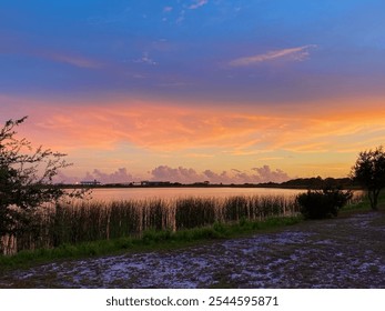 A vivid sunset over calm waters in Gulf Shores, Alabama, with vibrant orange and purple hues lighting up the sky, reflected in the water, surrounded by marshland. - Powered by Shutterstock