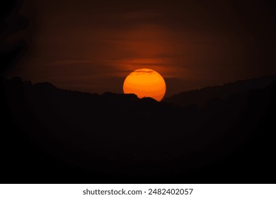 A vivid sunset over the hills, with the sun appearing as a large, glowing orb against a darkened sky. Silhouetted trees and mountains enhance the dramatic ambiance of the scene. - Powered by Shutterstock