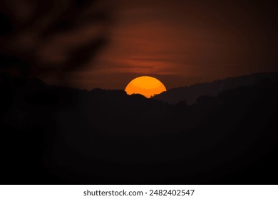 A vivid sunset over the hills, with the sun appearing as a large, glowing orb against a darkened sky. Silhouetted trees and mountains enhance the dramatic ambiance of the scene. - Powered by Shutterstock
