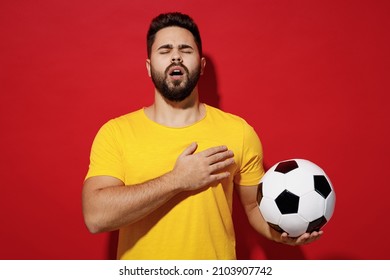 Vivid young bearded man football fan in yellow t-shirt cheer up support favorite team eyes closed hold soccer ball sing national country anthem isolated on plain dark red background studio portrait