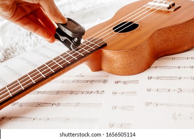 Vladivostok, Russia - 02.26.20: Tuning your guitar with a tuner at home. High-quality beautiful photo of a musical wooden instrument Ukulele with nylon strings on the background of musical notes.