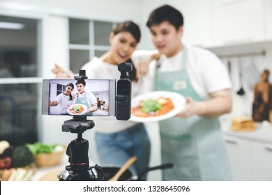 Vlogger And Blogger Freelance Job Concept, Young Asian Couple Cooking Together And Recording Live Video For Her Vlog And Social Media With Mobile Phone Camera And Tripod