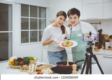 Vlogger And Blogger Freelance Job Concept, Young Asian Couple Cooking Together And Recording Live Video For Her Vlog And Social Media With Mobile Phone Camera And Tripod