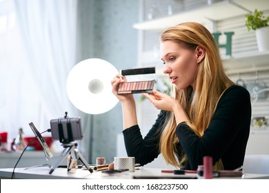 Vlogger female showing eye shadow palette. Beauty blogger woman filming daily make-up routine tutorial near camera on tripod. Influencer girl live streaming cosmetics product review in home studio.