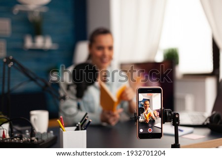 Vlogger holding book during podcast review on social media. Creative content creator influencer streaming live video, recording digital social media communication for her audience