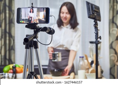 Vlogger Recording Content For The Baking Vlog Using A Phone On Tripod. Young Woman Filming Herself Preparing Cake In Kitchen.