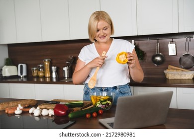 Vlogging blogging culinary vlogger. Caucasian young plus size plump woman cooking vegetable salad, preparing vegetarian vegan food meal, showing pepper in camera of laptop at home kitchen - Powered by Shutterstock