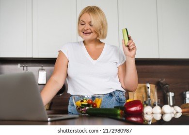 Vlogging blogging culinary vlogger. Plus size plump woman cooking vegetable salad, preparing vegetarian vegan food meal, showing cucumber in camera of laptop at home kitchen - Powered by Shutterstock