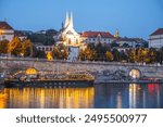 The Vltava River flows past the illuminated Emmaus Monastery in Prague, Czechia. The water reflects the lights of the city, creating a beautiful scene. A boat is docked near the riverbank.