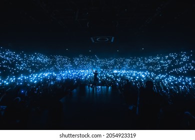 Vocalist in front of crowd on scene in stadium. Bright stage lighting, crowded dance floor. Phone lights at concert. Band blue silhouette crowd. People with cell phone lights.