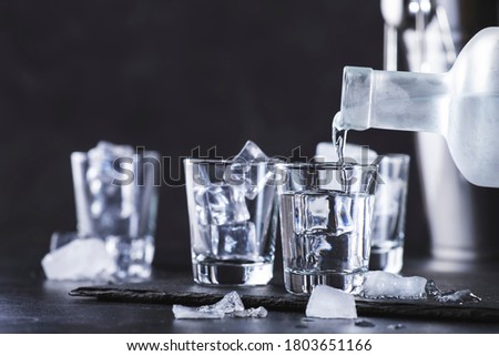 Vodka in shot glasses pouring out of the bottle on black stone background, iced strong drink in misted glass