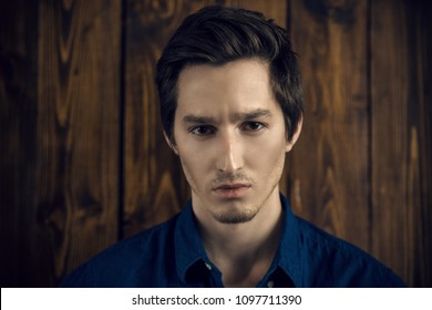 Vogue shot of a handsome male model in a denim shirt standing by a wooden wall. Men's beauty, fashion. 