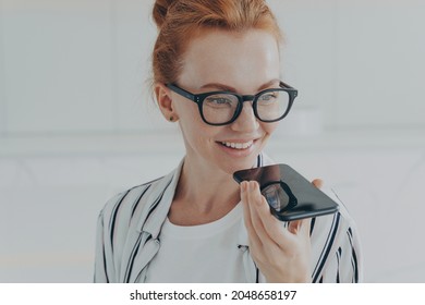 Voice search. Close up shot of redhead woman in transparent glasses speaks to virtual assistant holds mobile phone near mouth poses against white background smiles broadly. Technology communication - Powered by Shutterstock