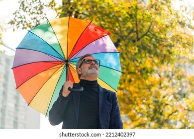 Volatile autumn weather. Lazy Sunday walk in nearby park. Medium closeup outdoor shot of elegant caucasian bearded man with black glasses holding big vivid rainbow umbrella and looking at the sky