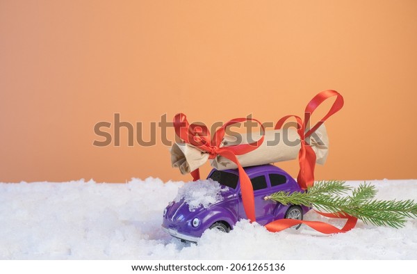 Volgograd, Russia - October 15, 2021:\
Christmas card concept. Christmas tiny car with gift boxes and a\
Christmas tree on an orange background with white\
snow.