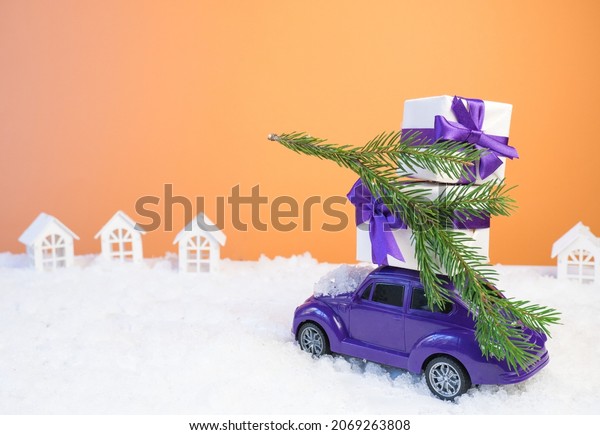 Volgograd, Russia - October 15, 2021:\
Christmas card concept. Christmas tiny car with gift boxes and a\
Christmas tree on an orange background with white\
snow