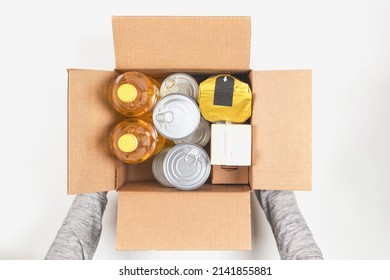 Volunteer Collecting Food Into Donation Box. Top View Woman Hands Holding Cardboard Box With Grocery Products On White Table. Donation, Charity, Food Bank, Help For Poor Families, Migrants, Refugees