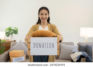 Volunteer Donation Clothes to Charity. Smiling Woman Holding Donation Box with Clothing in Living Room. Community Support and Generosity Concept - Powered by Shutterstock