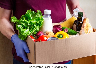 Volunteer In Gloves Holding Food In A Donation Cardboard Box With Various Food. Open Cardboard Box With Oil, Vegetables, Milk, Canned Food, Cereals And Pasta. Food Delivery Concept With Copy Space.