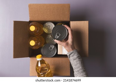 Volunteer Hands Collecting Food Into Donation Box. Top View To Cardboard Box With Grocery Products. Donation, Charity, Food Bank, Help For Poor Families, Migrants, Refugees