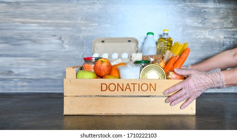 Volunteer Hands In Medical Gloves Holding Donation Box With Various Products For Poor People. Pandemic Coronavirus Food Shortages. Food Donations Or Delivery Concept. Free Space For Text Copyspace