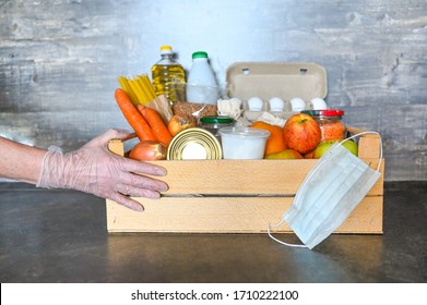 Volunteer Hands In Medical Gloves And Mask Holding Donation Box With Various Products For Poor. Pandemic Coronavirus Food Shortages. Food Donations Or Delivery Concept. Free Space For Text Copyspace