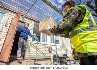 Volunteer In A Medical Mask And Gloves  Brings Food To Elderly Couple To Their Home. Volunteer Is Giving The Parcel To The Gray Haired Man On The Steps To The House. Outstretched Hands For Help.