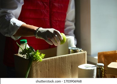 Volunteer In The Protective Medical Mask And Gloves Putting Food In Donation Box. Delivery Man Employee In Red Vest Packing Box With Food. Service Quarantine Pandemic Coronavirus.