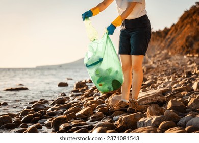 Volunteer in rubber gloves collects garbage on pebble wild beach and puts bottle in plastic bag. Copy space. Concept of Earth Day. - Powered by Shutterstock