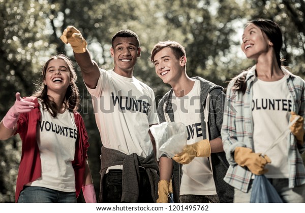 Volunteer\
sign. Beaming funny students wearing white shirt with volunteer\
sign feeling responsible while cleaning\
forest