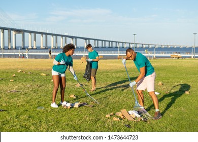 Volunteer Team Cleaning City Grass From Garbage. Men And Woman Working With Rakes And Picking Up Rubbish From Lawn. Trash Collection Concept