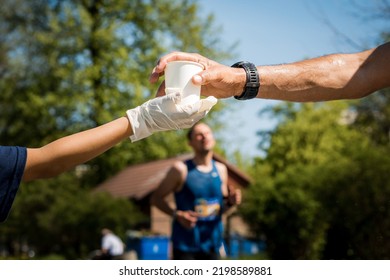 Volunteer In White Gloves Giving A Cup Of Water To Runner Of A Marathon. Refreshment Point. Runner Takes The Cup From Volunteers Hand. 