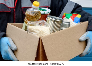 A Volunteer In A Winter Jacket Holds A Cardboard Box With Dry Cereals, Canned Food, Butter And Other Products. Help During A Pandemic. Donations Of Food Or The Concept Of Food Delivery.