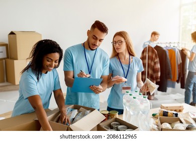 Volunteer Work. Young Arab Male Activist Checking Donation List And Writing In Clipboard, Volunteers Working In Charitable Organization, Packing Donations Boxes