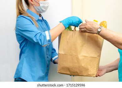 Volunteer Young Blonde Female In Blue Uniform And Medical Mask And Gloves Handing An Senior Woman A Paper Bag With Food. Donation, Support People In Quarantine, Coronavirus. Delivery Of Purchases.