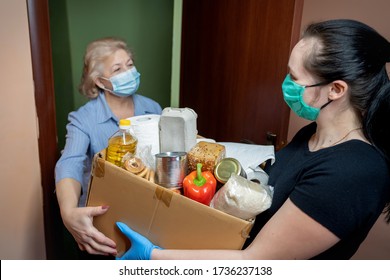 Volunteer Young Female In Medical Mask And Gloves Handing An Senior Woman A Box With Food. Donation, Support People In Quarantine, Coronavirus