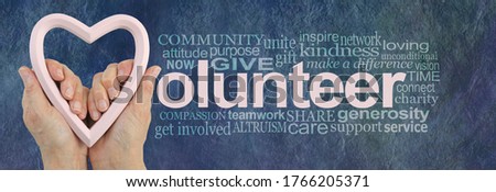 Volunteering is an act of LOVE Word Cloud - female hands holding a pale pink heart frame making the V of  VOLUNTEER beside a relevant word cloud on a blue  rustic stone effect background
