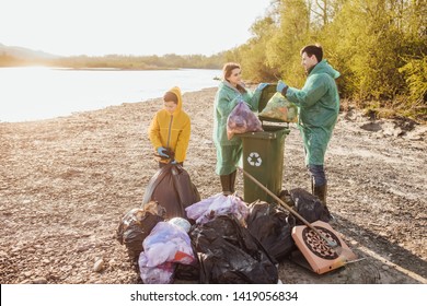 Volunteering, Charity, Cleaning, People And Ecology Concept - Group Of Happy  Family Volunteers With Garbage Bags Cleaning Area In Park Near Lake.