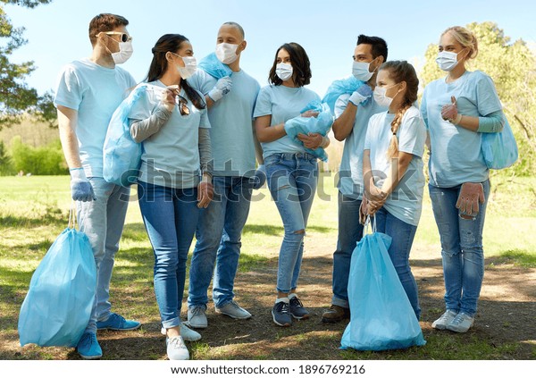 volunteering, health and\
ecology concept - group of volunteers wearing face protective\
medical mask for protection from virus disease with garbage bags\
talking in park