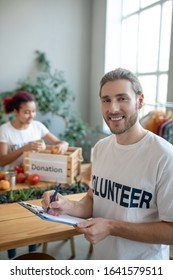 Volunteering. Young Bearded Smiling Kind Man In A White T-shirt With A Folder In His Hands, Standing Taking Notes, Girl Packing Fruits In A Box.