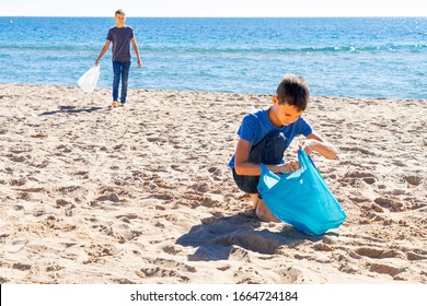 Volunteers Kids Cleaning Beach From Plastic. Boys Walking On The Beach And Picking Up Plastic Bottles Trash And Putting Into Plastic Bag For Recycle