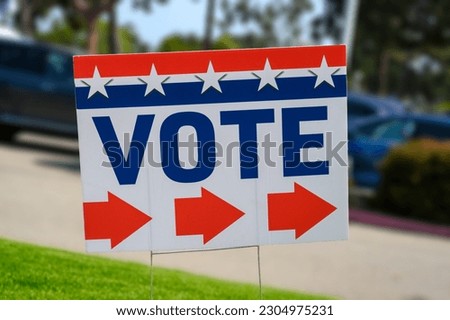 A VOTE sign at a polling place on green grass near a parking lot