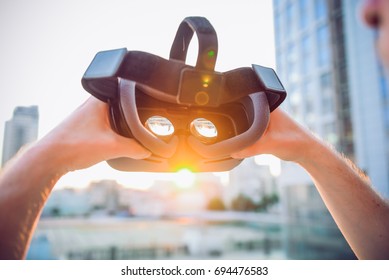 VR Goggles in male hands. Man ready to wear virtual reality goggles. The vr headset design is generic and no logos. Backward light, selective focus, close up. Sunset city landscape - Powered by Shutterstock