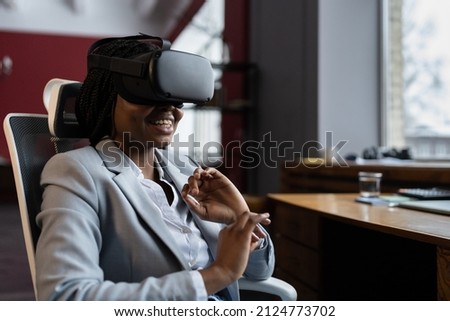 VR training in workplace. Smiling impressed black businesswoman sitting at office table using virtual reality glasses to reduce stress and relax at work. Power of augmented reality AR in business