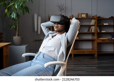 VR For Workplace Wellness. Amazed African American Female Employee In Virtual Reality Goggles Resting At In Relaxing Virtual Environment, Sitting On Office Chair With Hands Behind Head. AR In Business