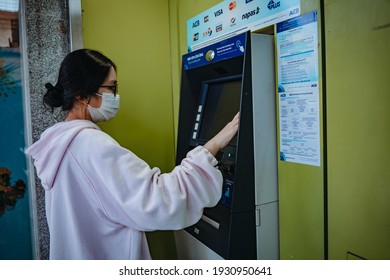 Vung Tau, VIETNAM - 5 MAR 2021: Close-up of woman (wearing medical mask and pink hoodie), using credit card to withdrawing money from ATM machine. Finance and business concept. Selective focus.
