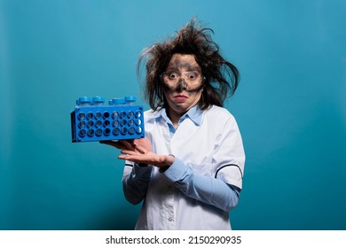 Wacky mad chemist with messy hair and dirty face holding plastic test tubes on rack while looking at camera on blue background. Lunatic biochemistry expert having stand with test flasks. Studio shot.