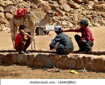 Wadi Rum, Jordan - May 2019: Poorly Dressed Children Sitting On A Rock Water Trench In The Wadi Rum Desert, With A Camel And A Pick-up Truck In The Background. 